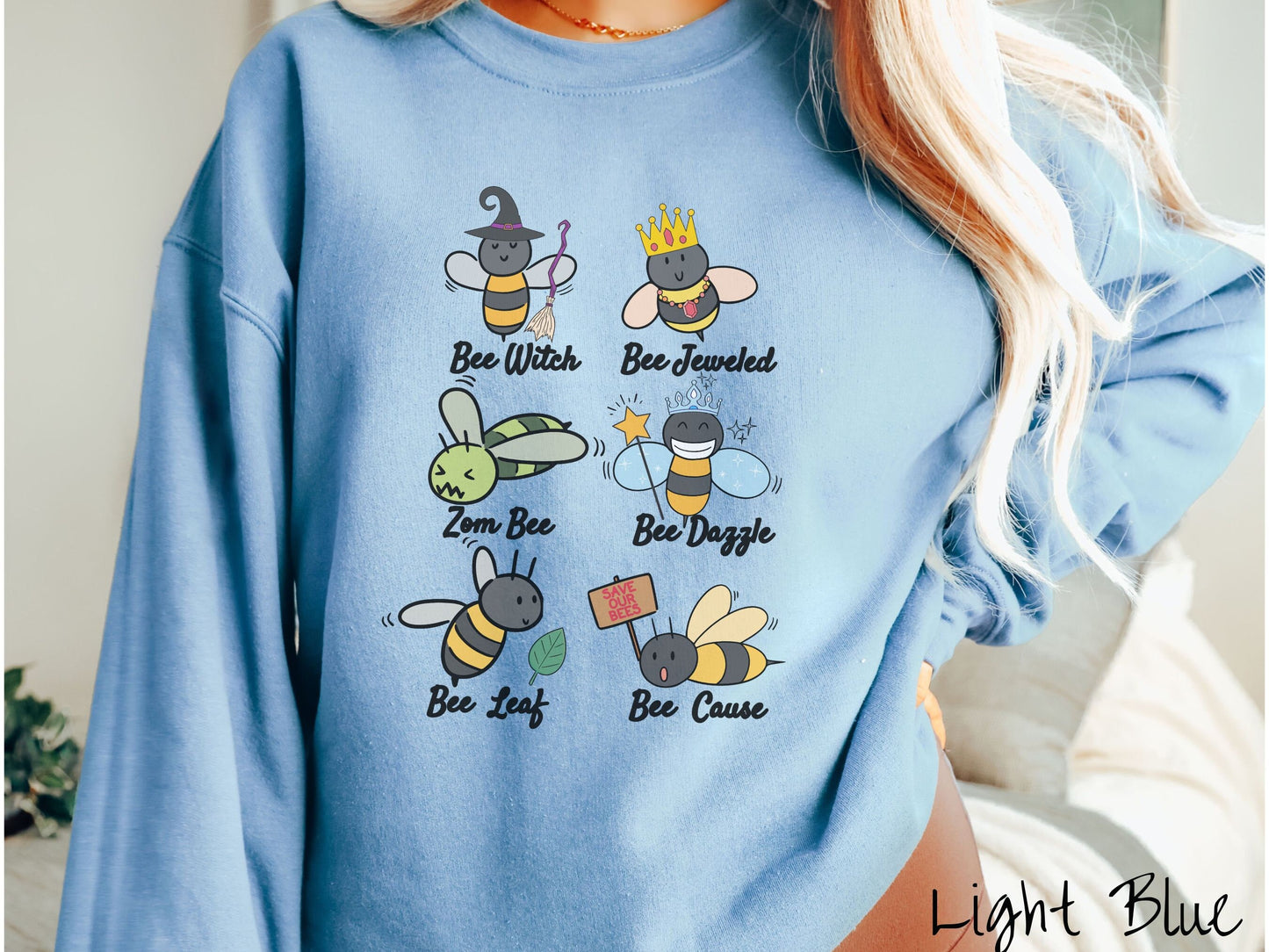 A woman wearing a cute, vintage light blue colored comfy sweatshirt that has six puns that play on honey bees accompanied with bee drawings. The puns are Bee Witch, Bee Jeweled, Zom Bee, Bee Dazzle, Bee Leaf, and Bee Cause.