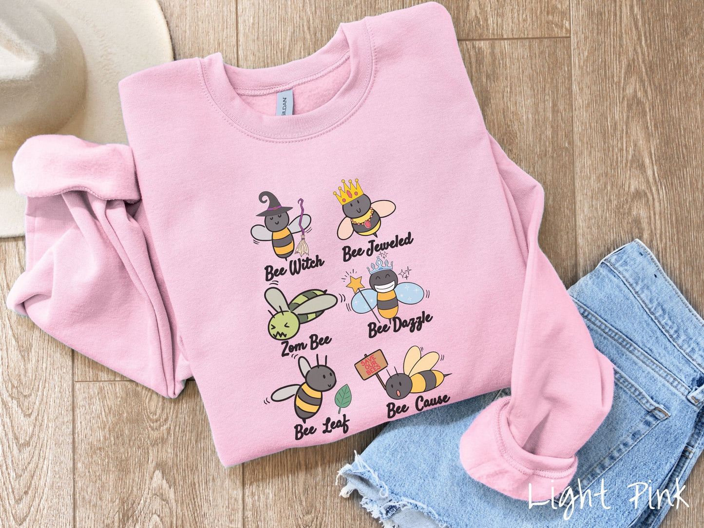 A cute, vintage light pink colored comfy sweatshirt that has six puns that play on honey bees accompanied with bee drawings. The puns are Bee Witch, Bee Jeweled, Zom Bee, Bee Dazzle, Bee Leaf, and Bee Cause.