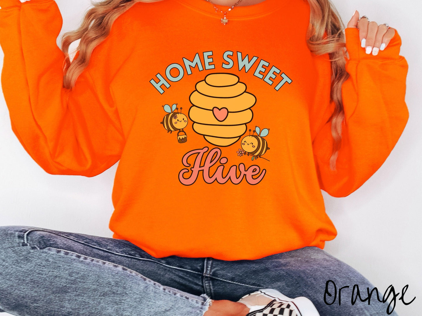 A woman wearing a cute, vintage orange colored comfy sweatshirt with the text Home Sweet Hive in cyan and red font. In between the words are a yellow bee hive with a red heart on it and smiling honey bees flying around holding flowers.