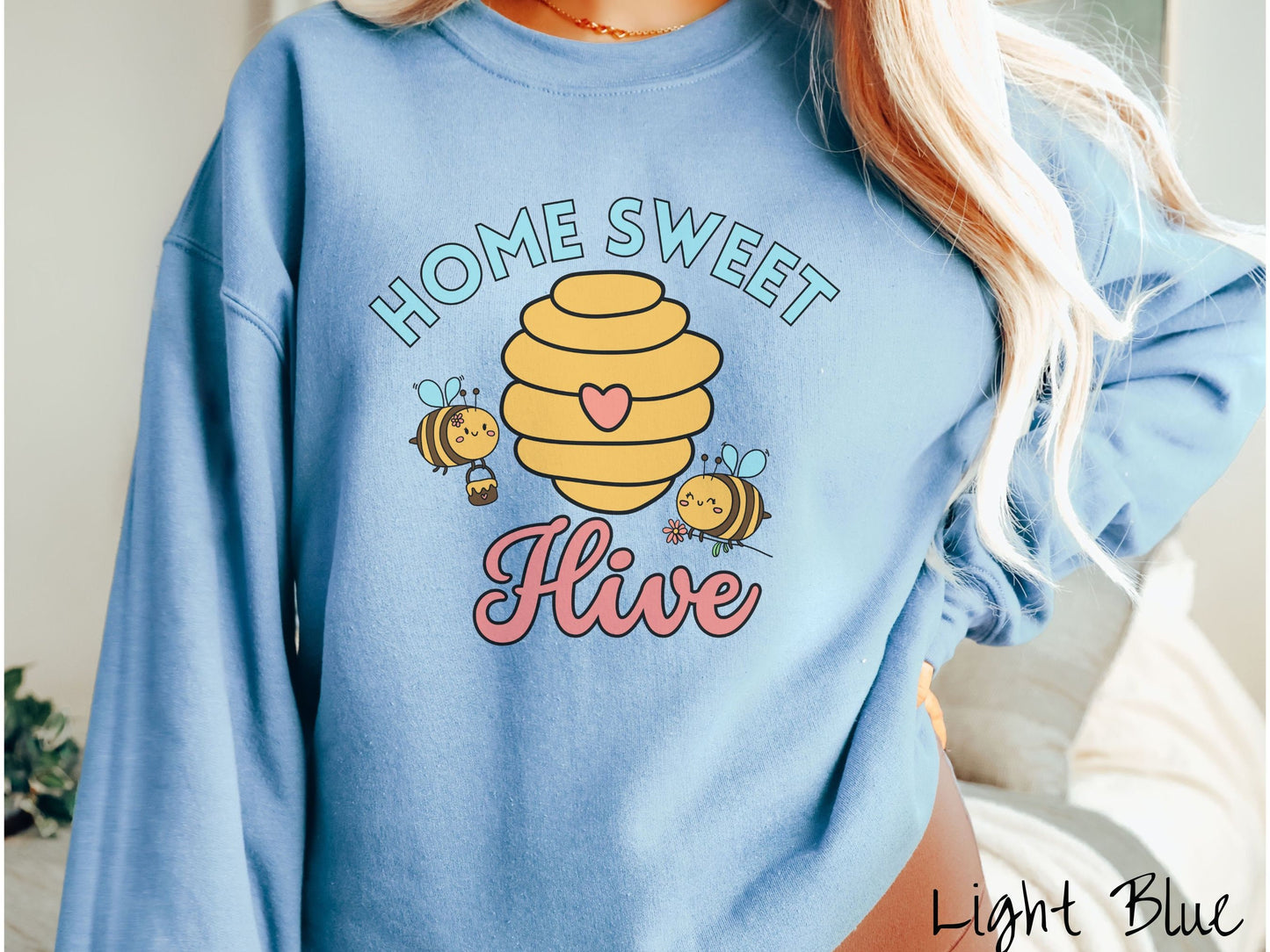 A woman wearing a cute, vintage light blue colored comfy sweatshirt with the text Home Sweet Hive in cyan and red font. In between the words are a yellow bee hive with a red heart on it and smiling honey bees flying around holding flowers.
