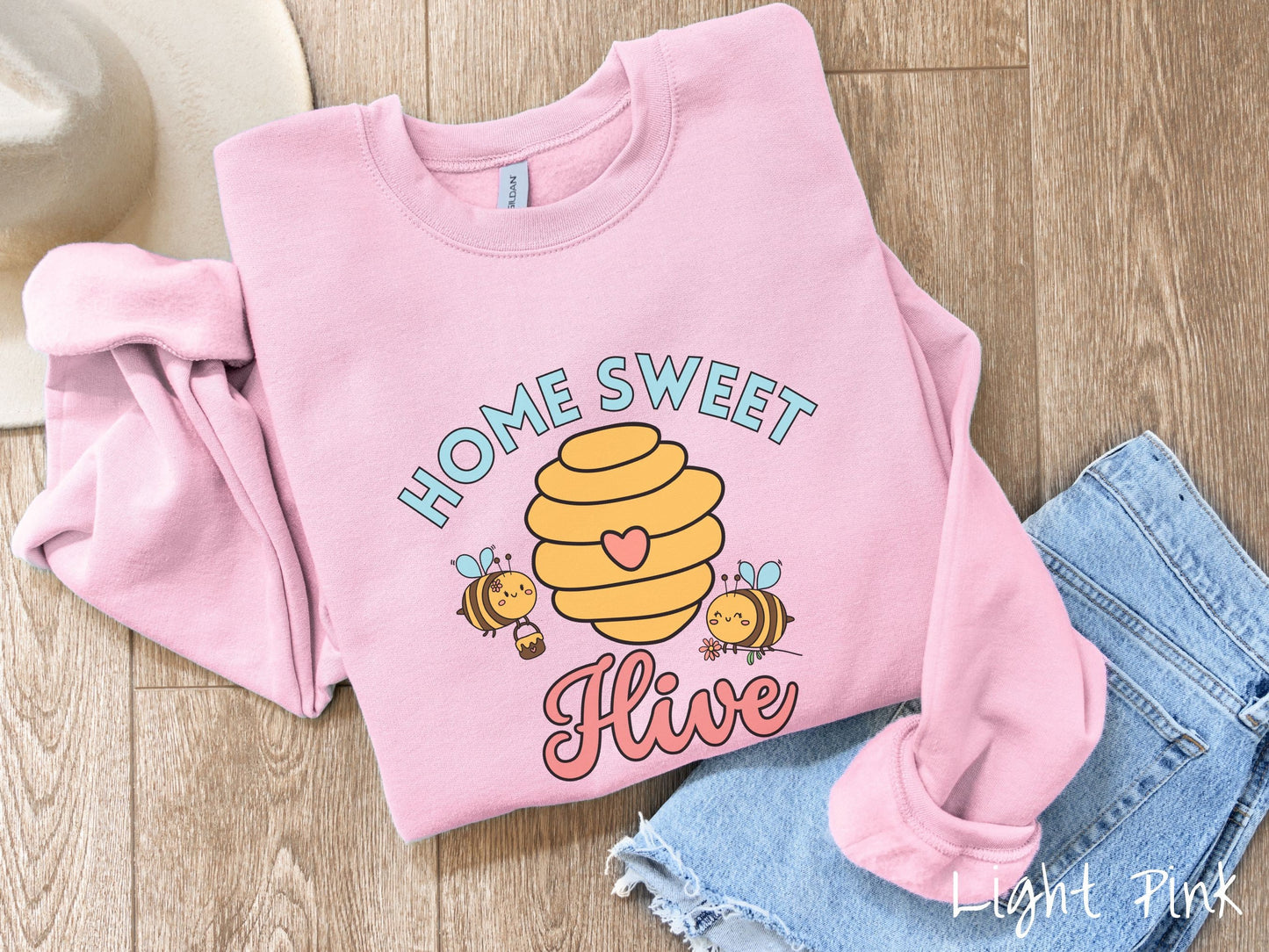 A cute, vintage light pink colored comfy sweatshirt with the text Home Sweet Hive in cyan and red font. In between the words are a yellow bee hive with a red heart on it and smiling honey bees flying around holding flowers.