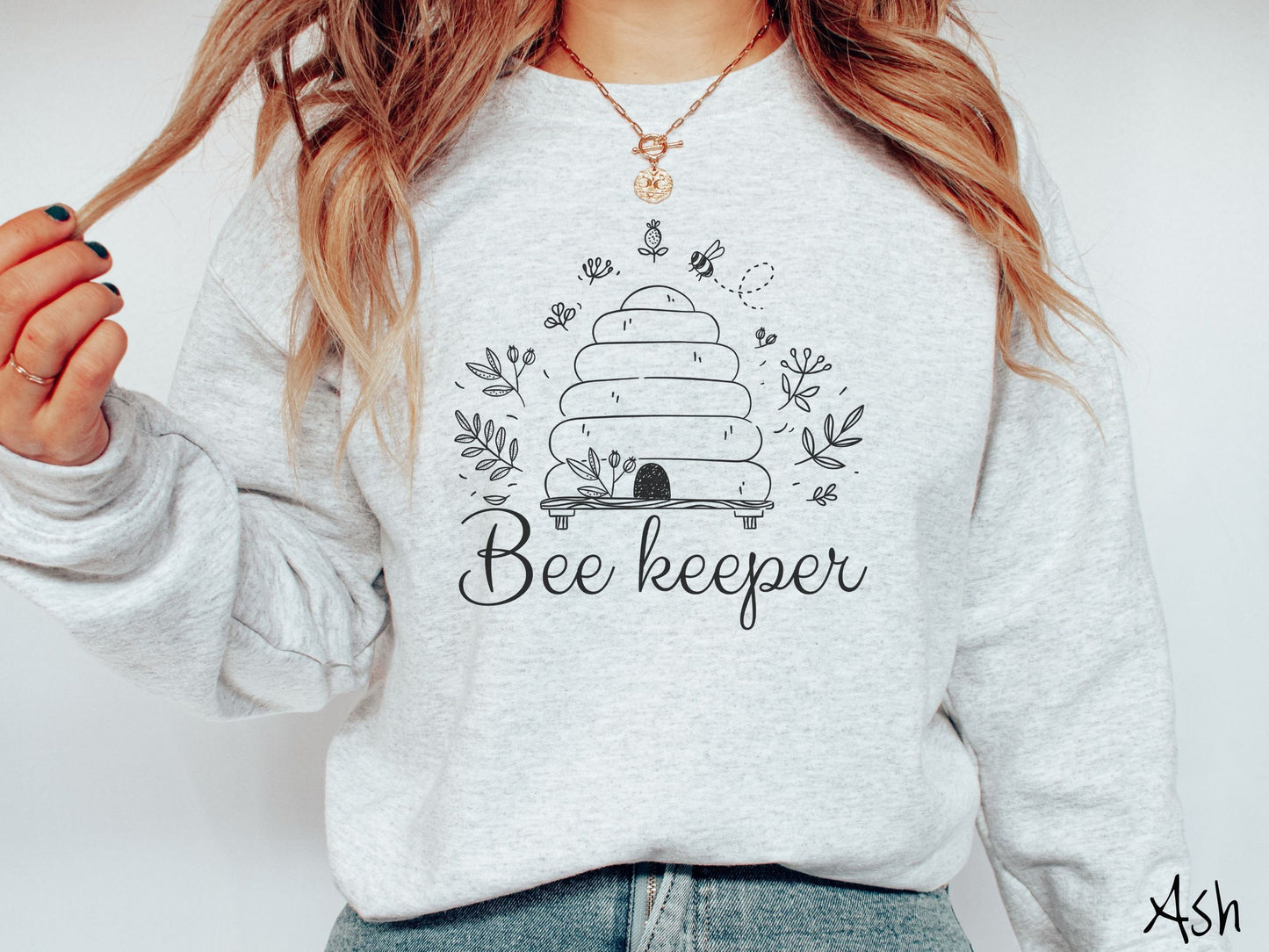 A woman wearing a cute, vintage ash colored comfy sweatshirt with the text Beekeeper across the front in black, cursive font. Above this is a black-outlined bee hive with bees buzzing around it and there are leaves in the air.