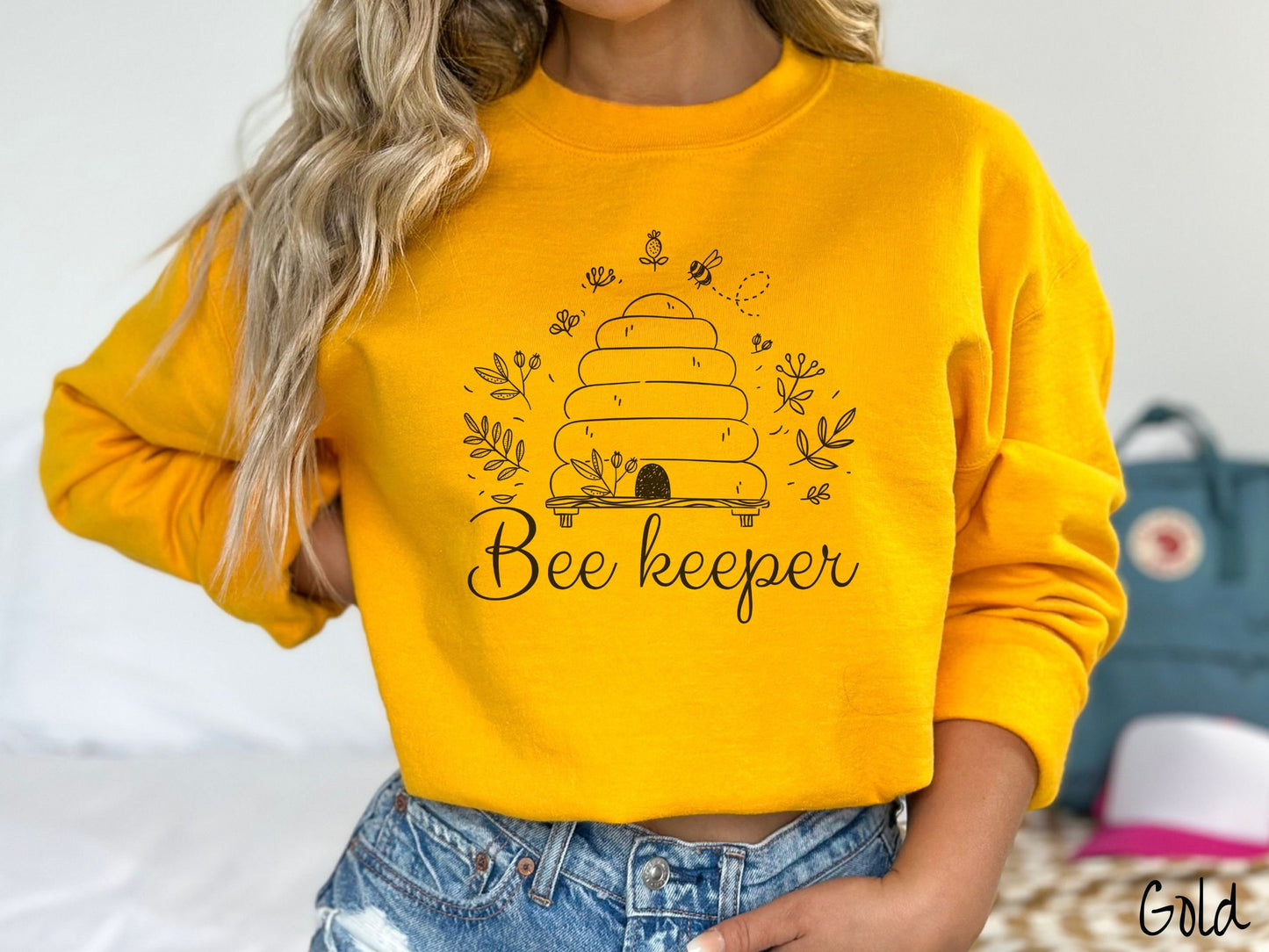 A woman wearing a cute, vintage gold colored comfy sweatshirt with the text Beekeeper across the front in black, cursive font. Above this is a black-outlined bee hive with bees buzzing around it and there are leaves in the air.