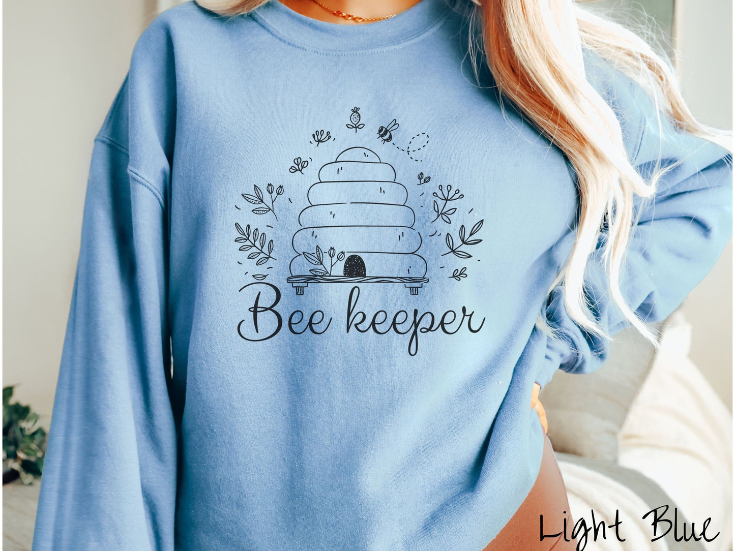 A woman wearing a cute, vintage light blue colored comfy sweatshirt with the text Beekeeper across the front in black, cursive font. Above this is a black-outlined bee hive with bees buzzing around it and there are leaves in the air.