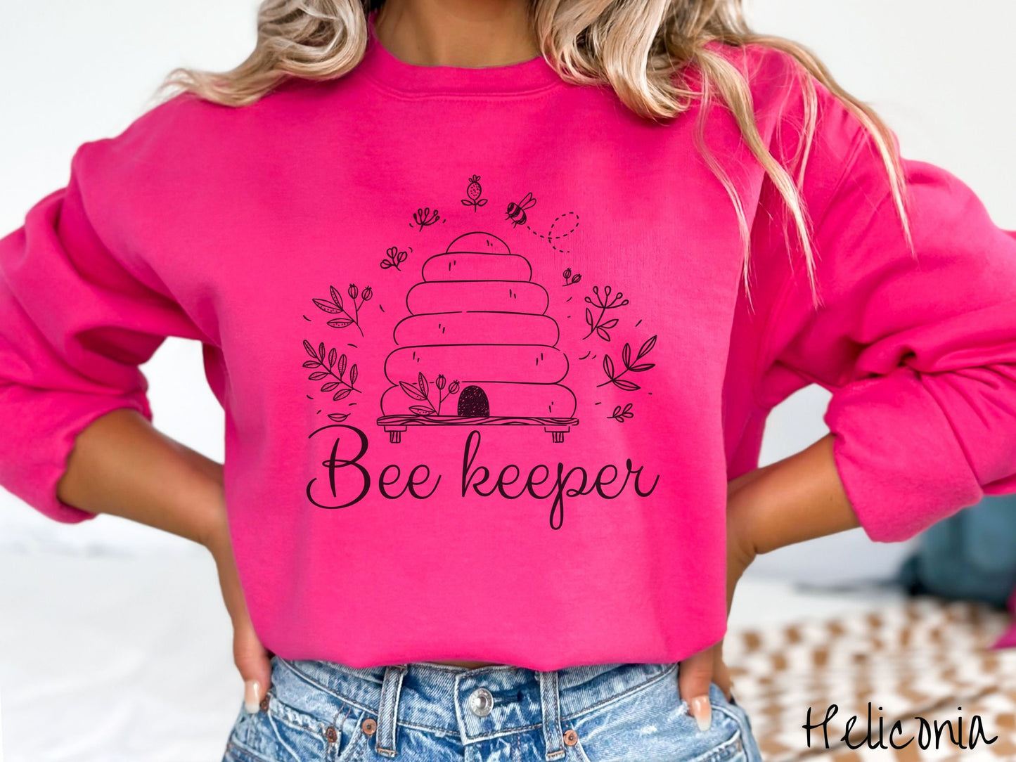 A woman wearing a cute, vintage heliconia colored comfy sweatshirt with the text Beekeeper across the front in black, cursive font. Above this is a black-outlined bee hive with bees buzzing around it and there are leaves in the air.