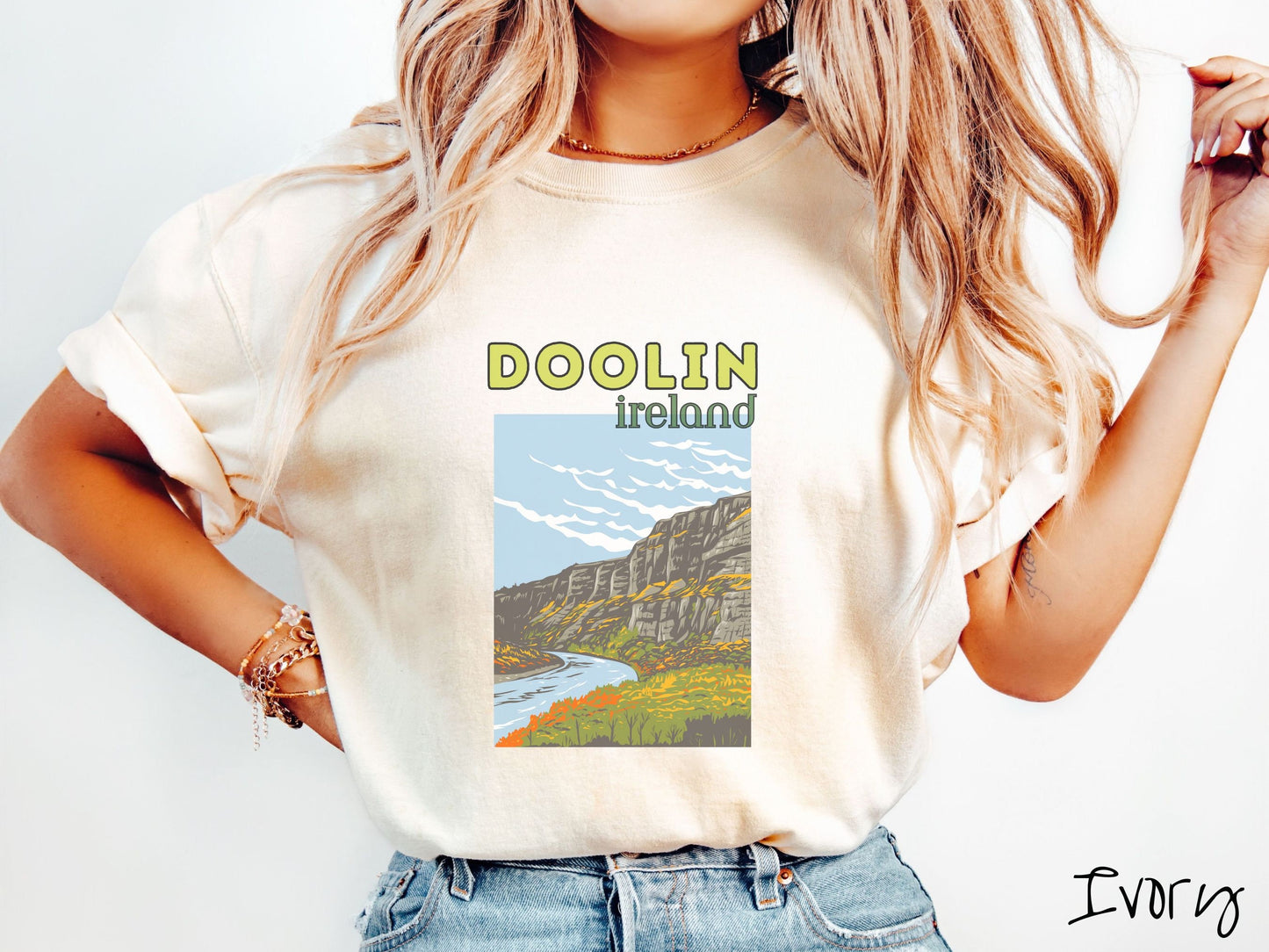 A woman wearing a vintage, ivory colored shirt with the text Doolin Ireland in yellow and green font, respectively, with a picture of an Ireland landscape below, steep cliffs overlooking a rushing river and green meadows in the Irish highlands.
