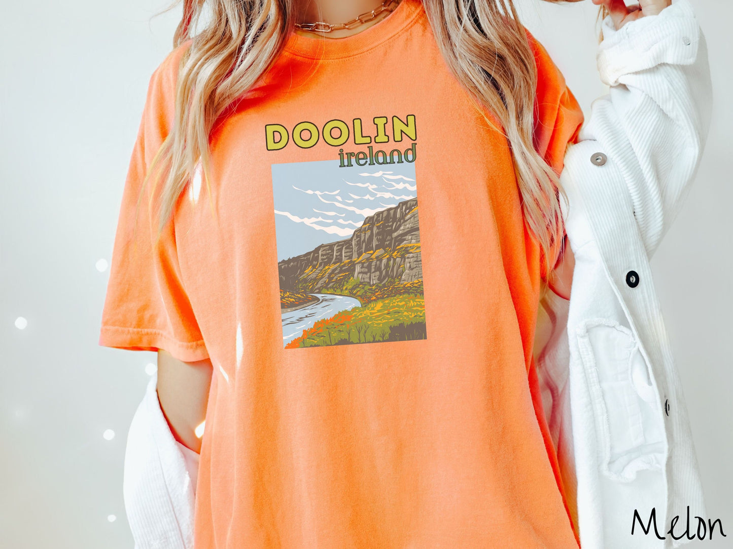 A woman wearing a vintage, melon colored shirt with the text Doolin Ireland in yellow and green font, respectively, with a picture of an Ireland landscape below, steep cliffs overlooking a rushing river and green meadows in the Irish highlands.