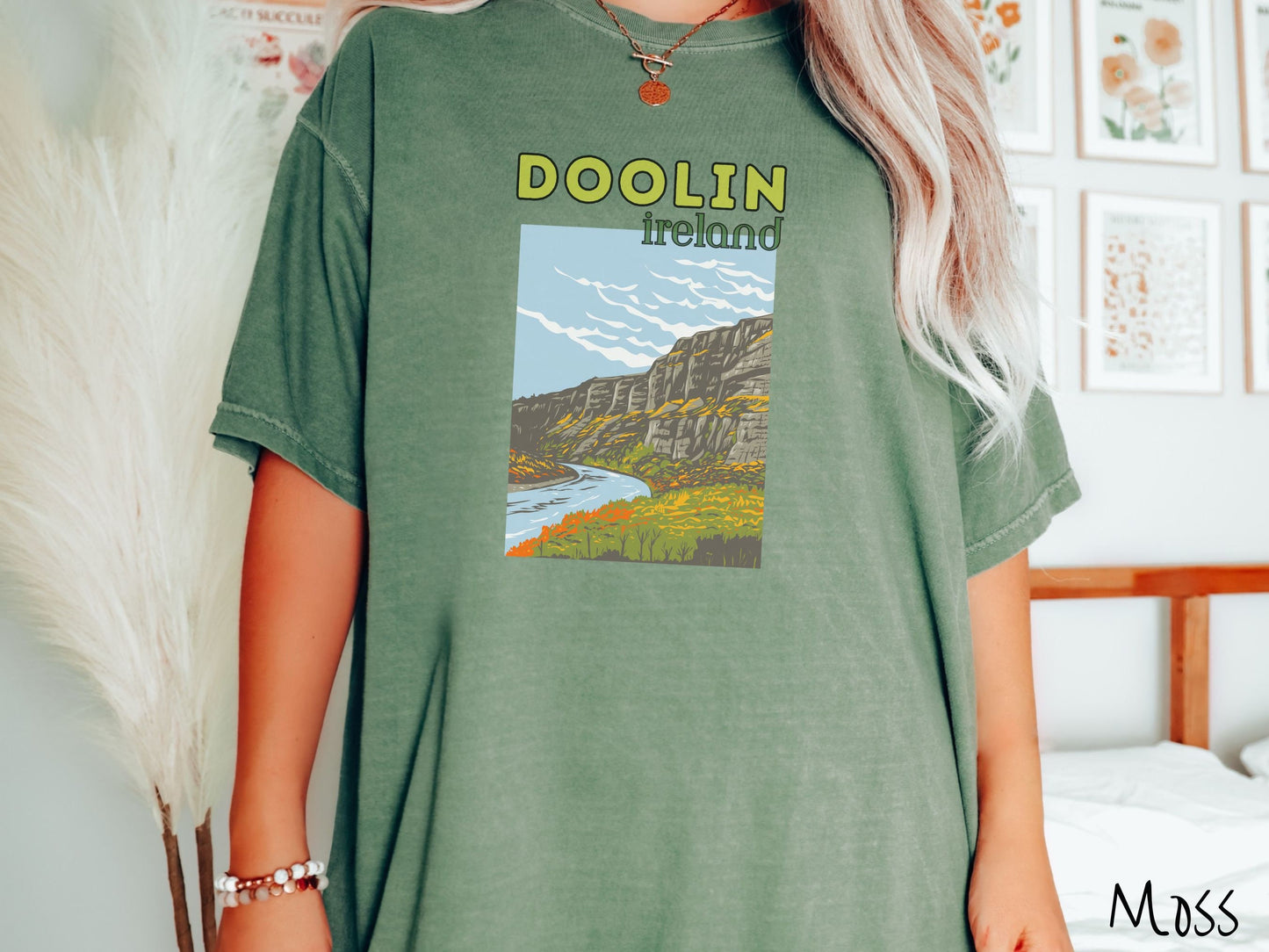 A woman wearing a vintage, moss colored shirt with the text Doolin Ireland in yellow and green font, respectively, with a picture of an Ireland landscape below, steep cliffs overlooking a rushing river and green meadows in the Irish highlands.