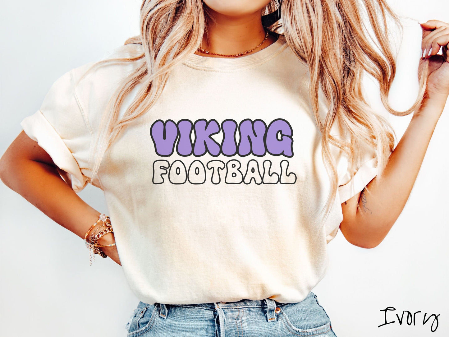 A woman wearing a cute, vintage ivory colored Comfort Colors shirt with the word Viking across the front in purple, uppercase font. Below that is the word Football in white, uppercase font.