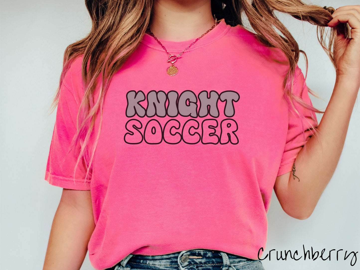 A woman wearing a cute, vintage crunchberry colored Comfort Colors shirt with the word Knight across the front in gray, uppercase font. Below that is the word Softball in pink, uppercase font.