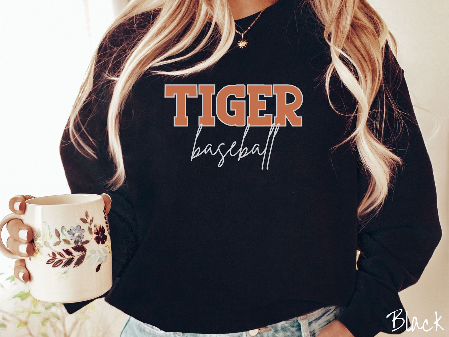 A woman wearing a cute, vintage black colored sweatshirt with the word Tiger in uppercase, orange font across the front and the word baseball below that in white, undercase cursive writing.