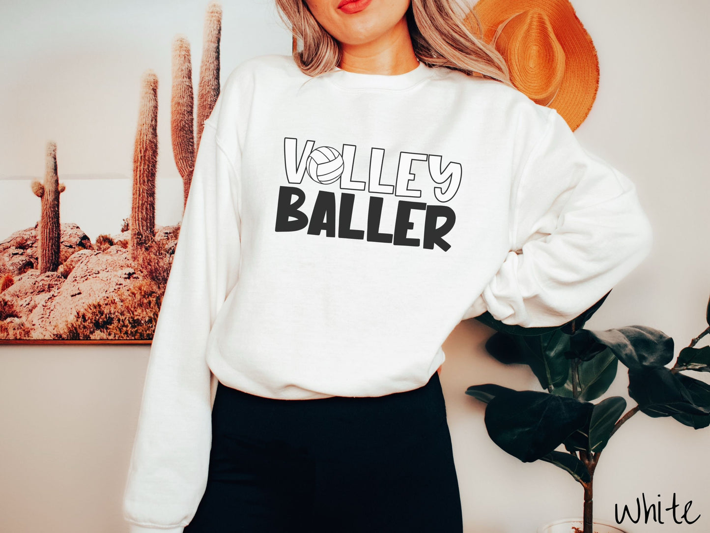 A woman wearing a cute, vintage white colored sweatshirt with the word Volley across the front in white, uppercase font. The O in Volley is a white volleyball. Below that is the word Baller in black, uppercase font.