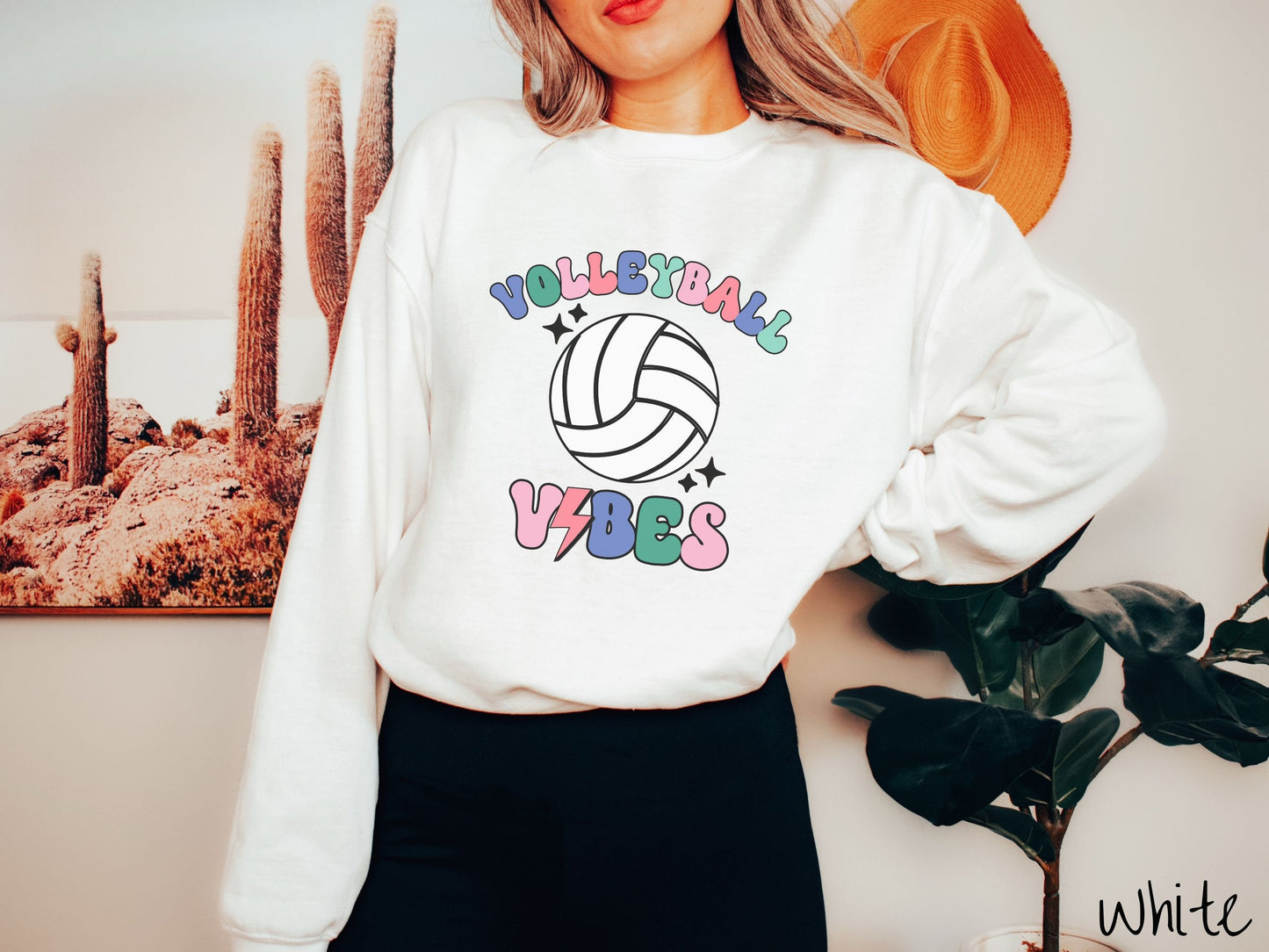 A woman wearing a cute, vintage white colored sweatshirt with the word Volleyball across the front in colorful, uppercase fonts. Below that is a white volleyball and below that is the word vibes in colorful, uppercase fonts.