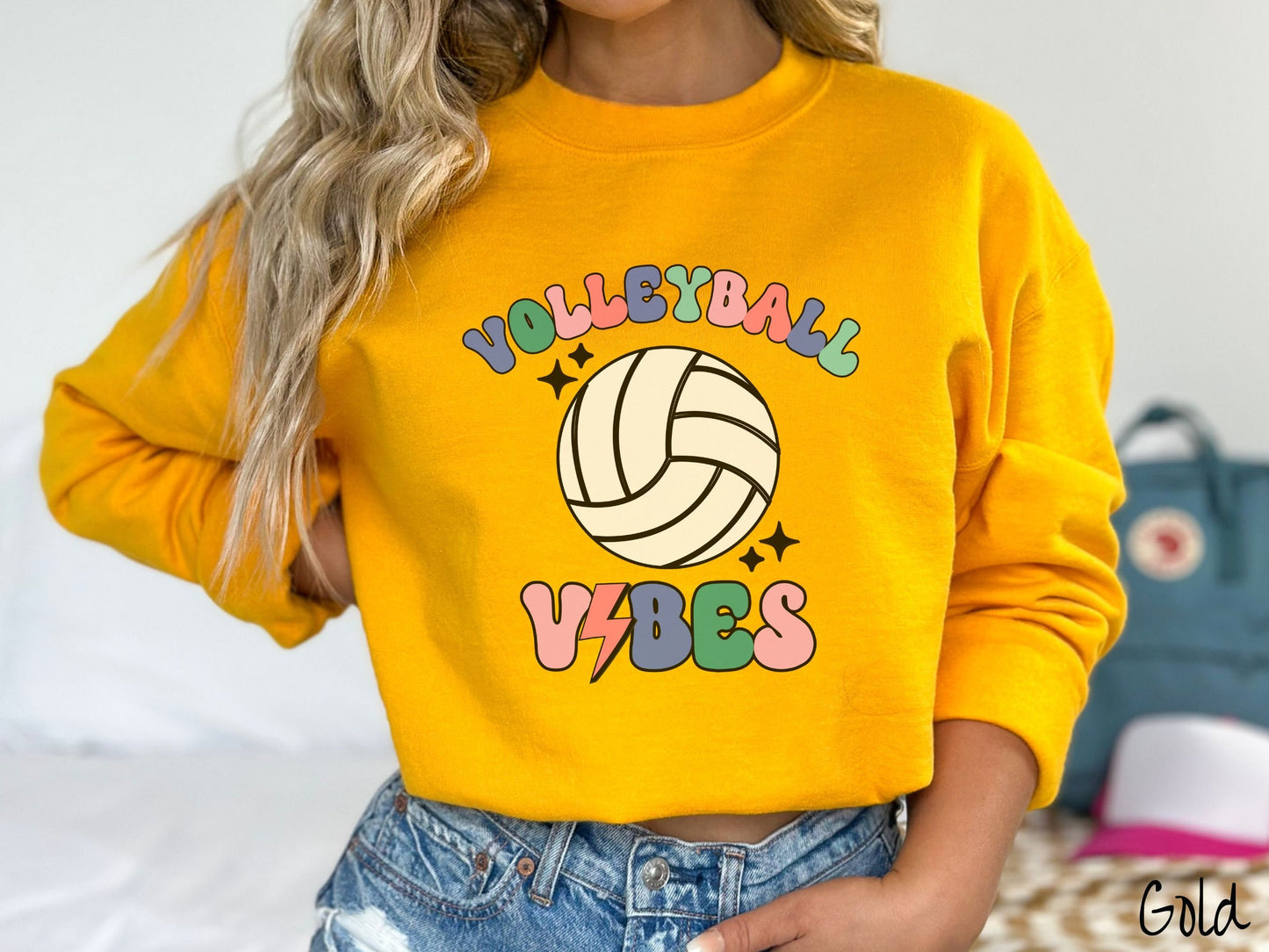 A woman wearing a cute, vintage gold colored sweatshirt with the word Volleyball across the front in colorful, uppercase fonts. Below that is a white volleyball and below that is the word vibes in colorful, uppercase fonts.