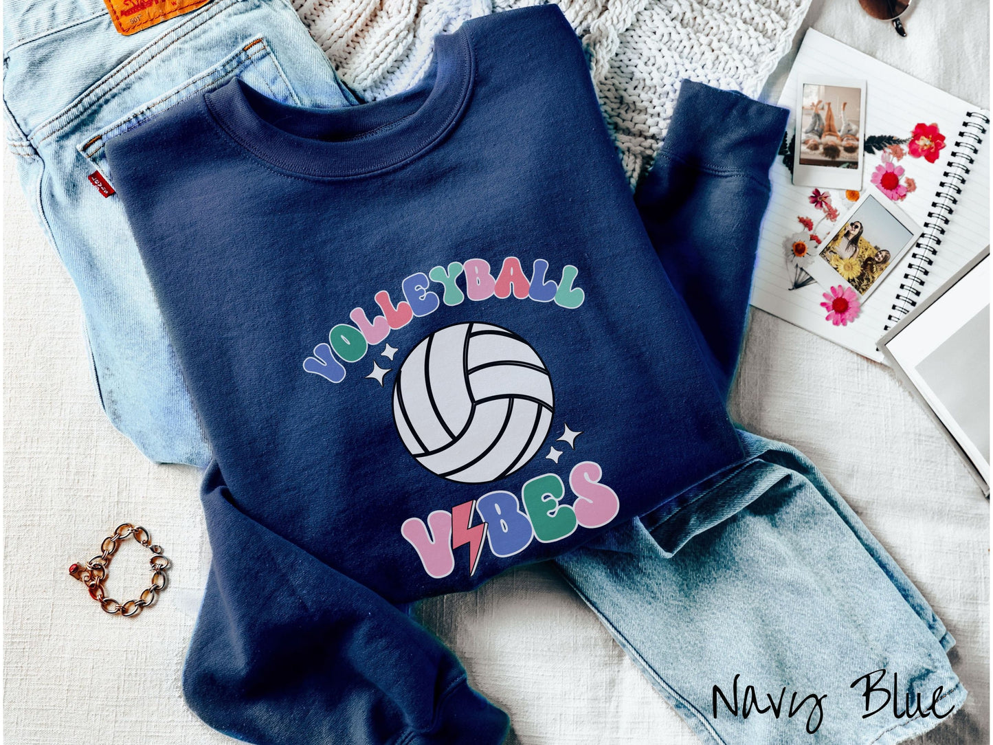 A woman wearing a cute, vintage navy blue colored sweatshirt with the word Volleyball across the front in colorful, uppercase fonts. Below that is a white volleyball and below that is the word vibes in colorful, uppercase fonts.