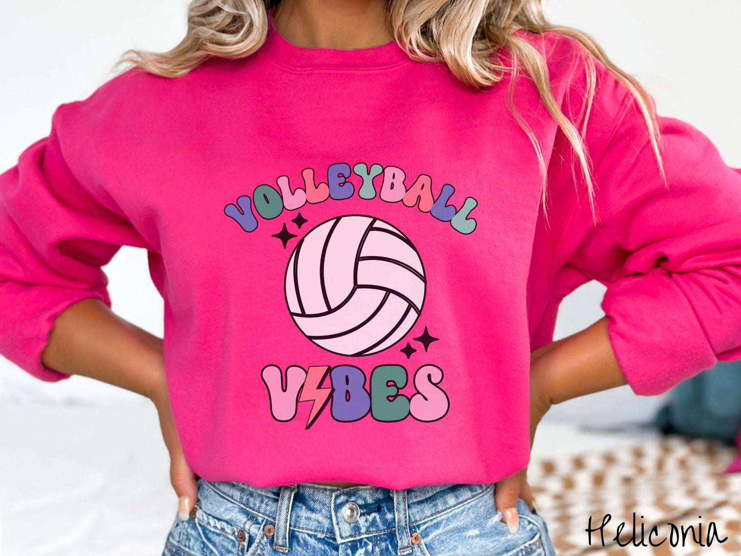 A woman wearing a cute, vintage heliconia colored sweatshirt with the word Volleyball across the front in colorful, uppercase fonts. Below that is a white volleyball and below that is the word vibes in colorful, uppercase fonts.