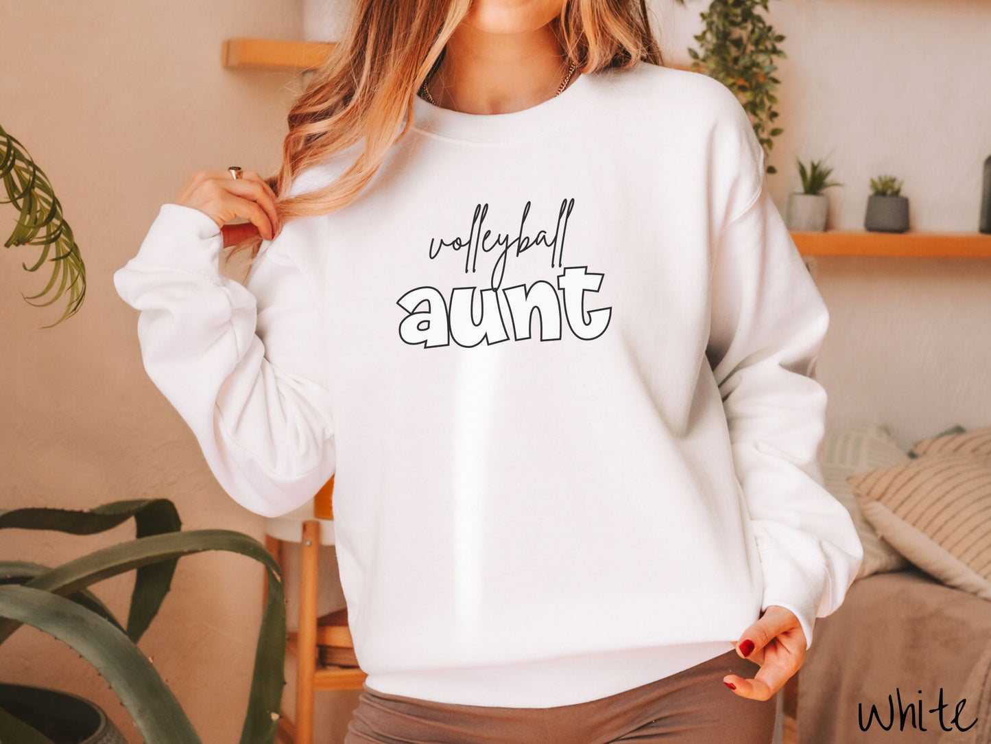 A woman wearing a cute, vintage white colored comfy sweatshirt with the word volleyball across the front in black, undercase cursive writing. Below that is the word aunt in large, white font.