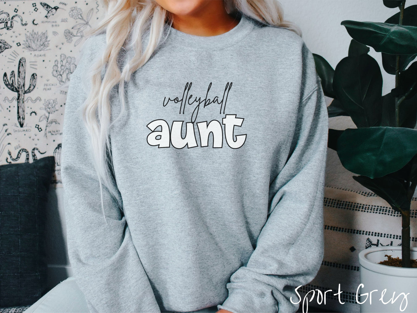 A woman wearing a cute, vintage sport grey colored comfy sweatshirt with the word volleyball across the front in black, undercase cursive writing. Below that is the word aunt in large, white font.