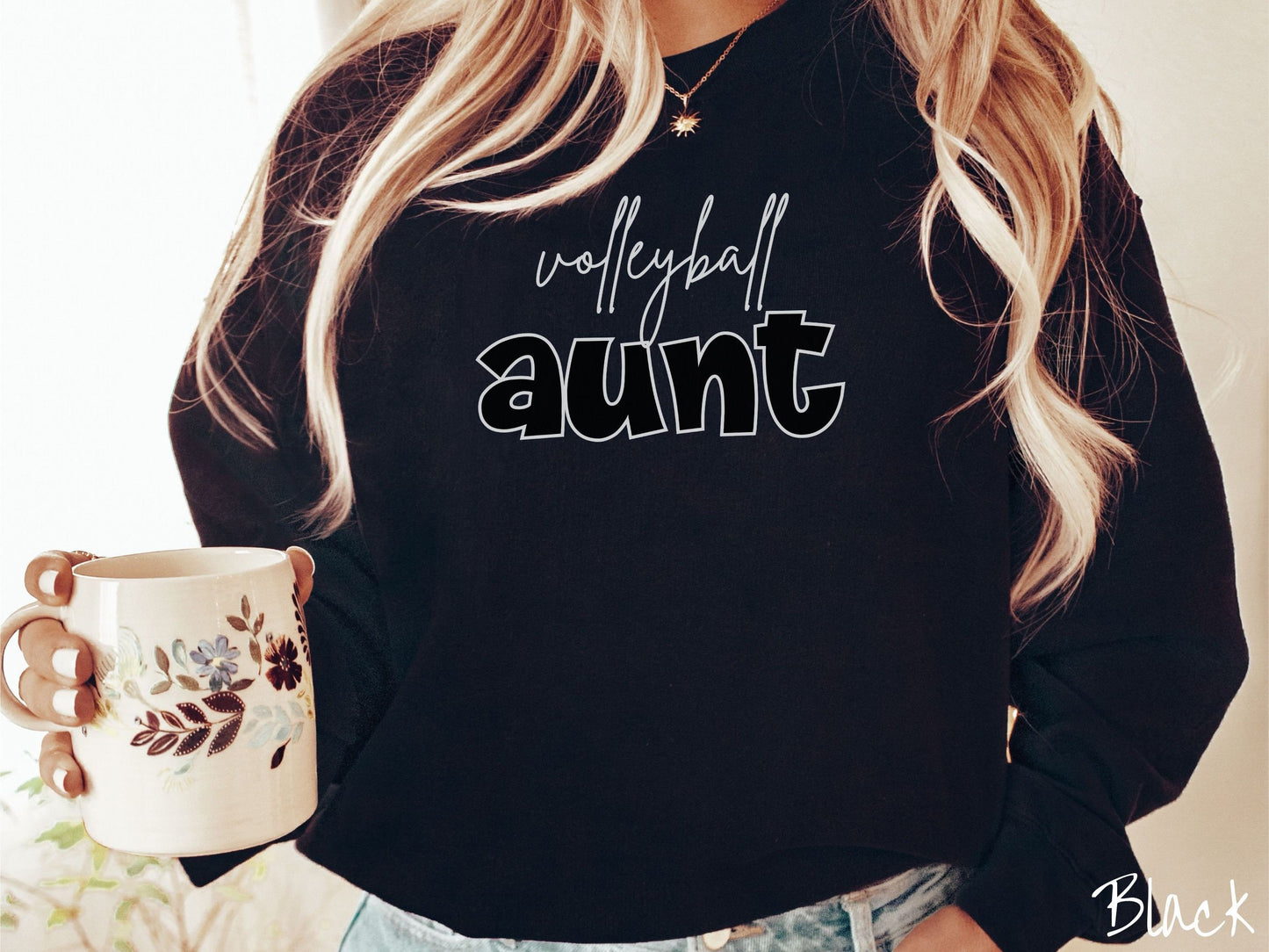 A woman wearing a cute, vintage black colored comfy sweatshirt with the word volleyball across the front in black, undercase cursive writing. Below that is the word aunt in large, white font.