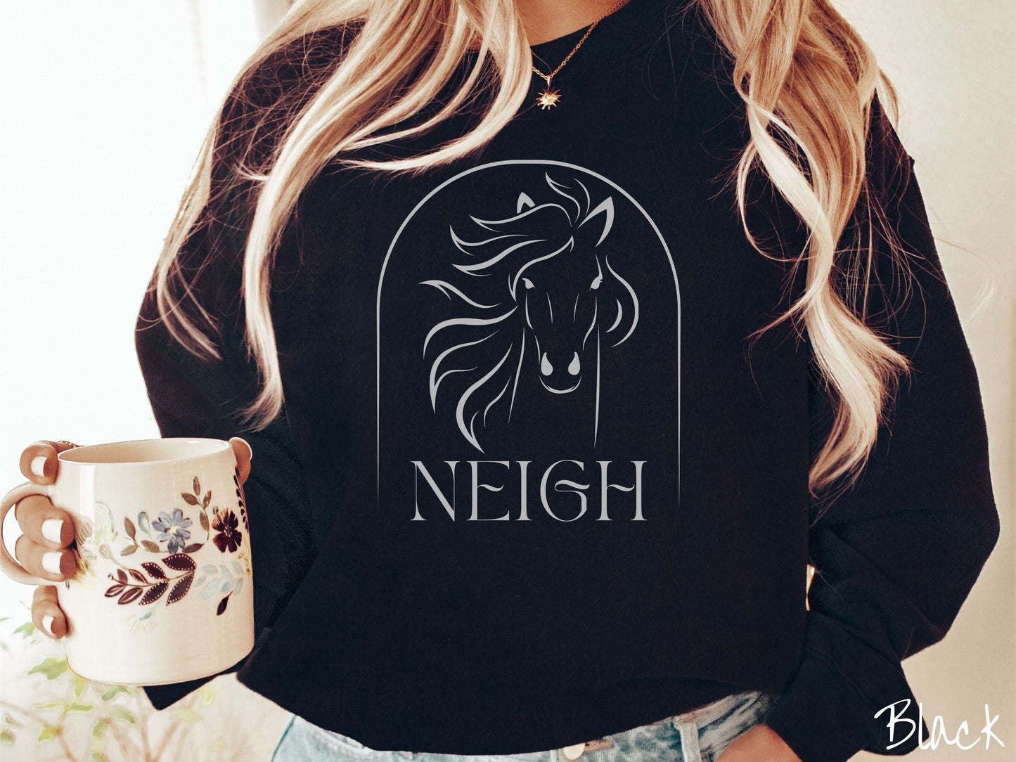 A woman wearing a cute, vintage black colored comfy sweatshirt with a white silhouette outline of a horse facing forward, its hair blowing to the side. Below that is the word Neigh in white, cursive font.