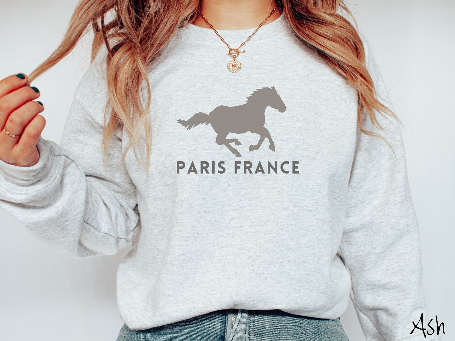 A woman wearing a cute, vintage ash colored comfy sweatshirt with a gray silhouette of a running horse across the font. Below it are the words Paris France in gray, uppercase font.
