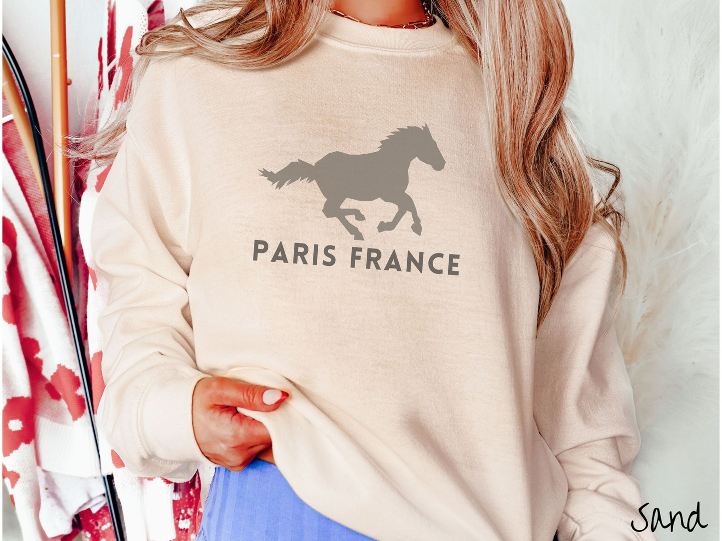 A woman wearing a cute, vintage sand colored comfy sweatshirt with a gray silhouette of a running horse across the font. Below it are the words Paris France in gray, uppercase font.