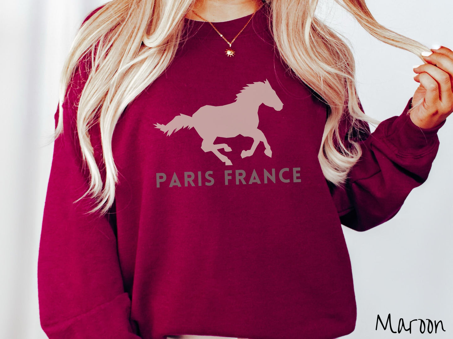 A woman wearing a cute, vintage maroon colored comfy sweatshirt with a gray silhouette of a running horse across the font. Below it are the words Paris France in gray, uppercase font.