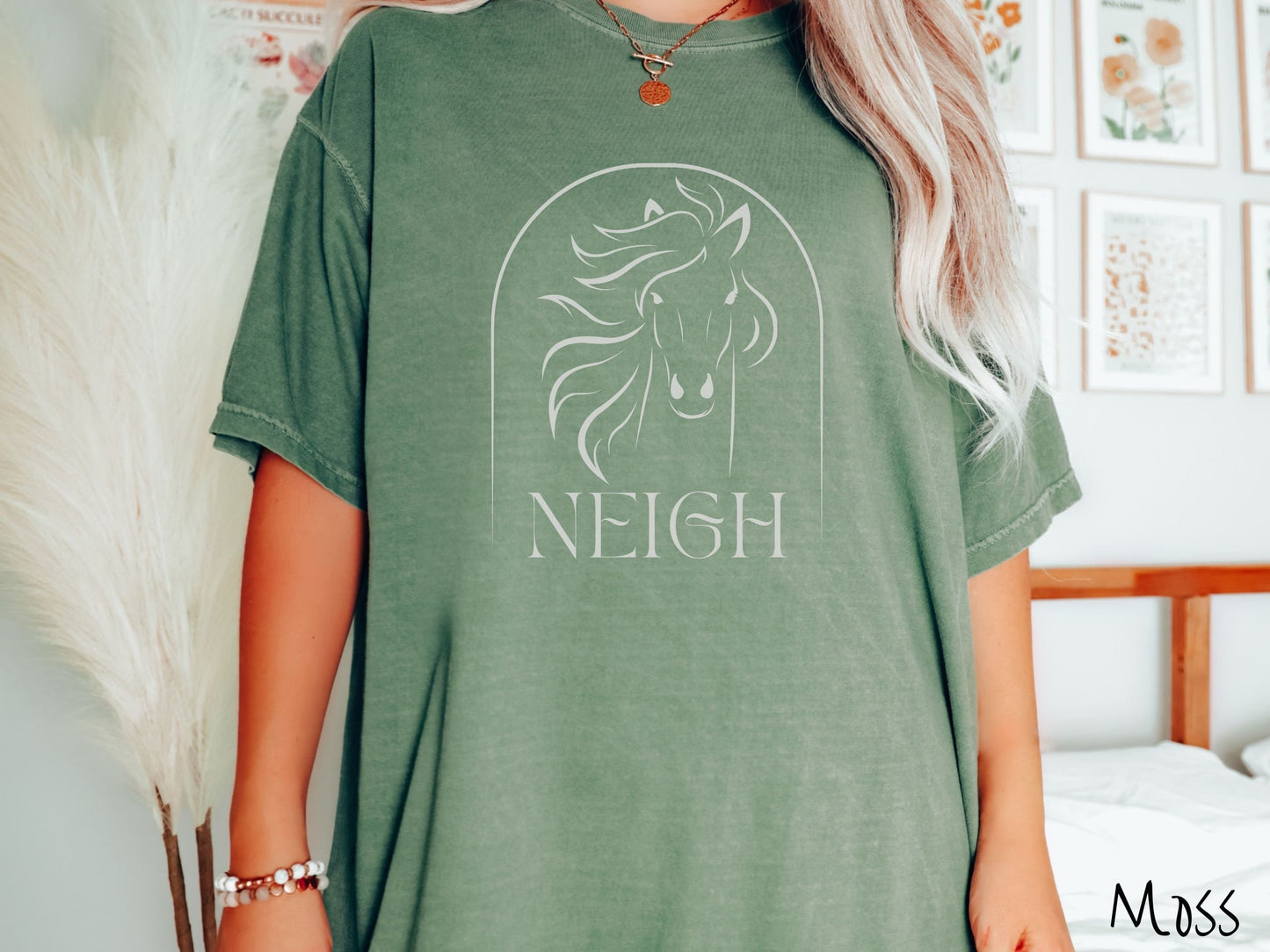 A woman wearing a cute, vintage moss colored Comfort Colors shirt with a white silhouette outline of a horse facing forward, its hair blowing to the side. Below that is the word Neigh in white, cursive font.