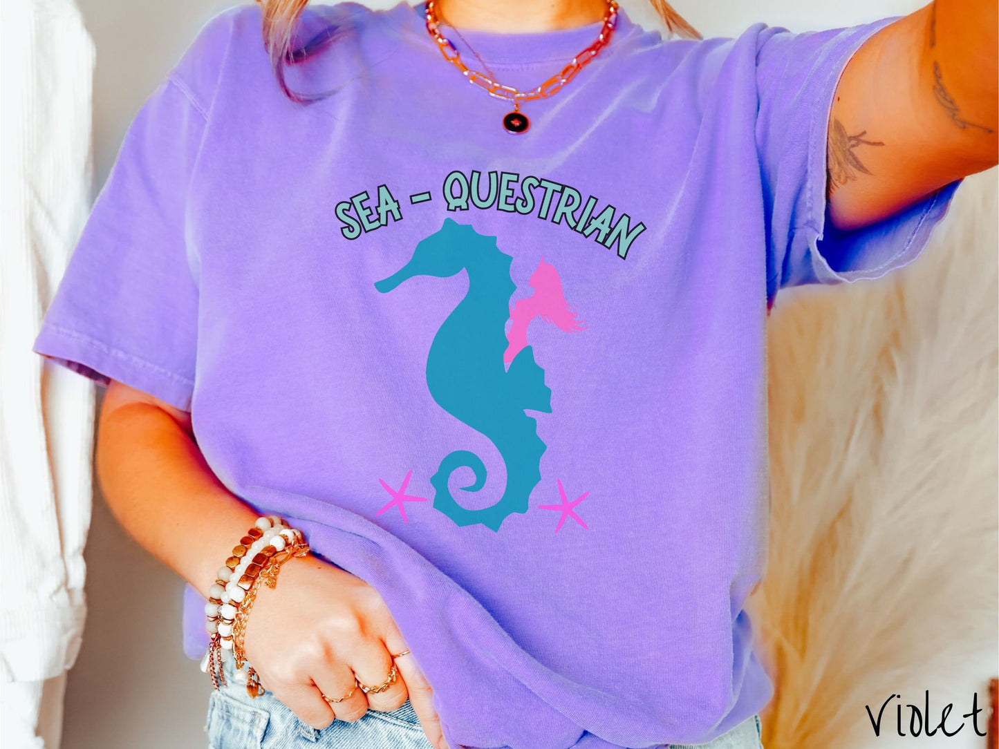 A woman wearing a cute, vintage violet colored Comfort Colors shirt with text Sea-Questrian in seafoam colored font across the top. Below this is a pink mermaid with flowing hair riding on the back of a turquoise seahorse with pink starfish below.