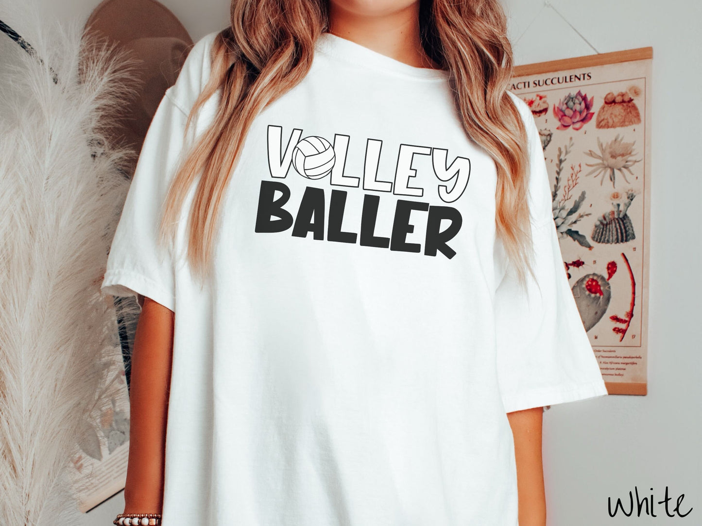 A woman wearing a cute, vintage white colored Comfort Colors shirt with the word Volley across the front in white, uppercase font. The O in Volley is a white volleyball. Below that is the word Baller in black, uppercase font.