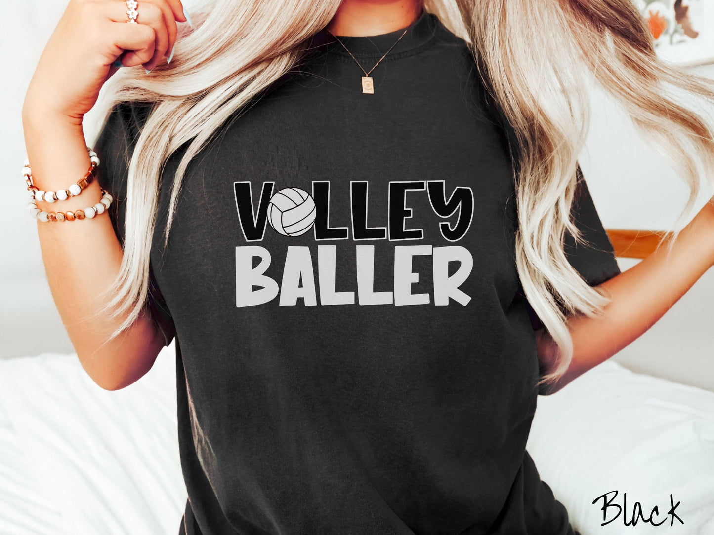 A woman wearing a cute, vintage black colored Comfort Colors shirt with the word Volley across the front in black, uppercase font. The O in Volley is a white volleyball. Below that is the word Baller in white, uppercase font.