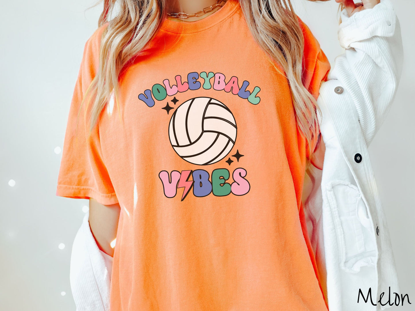 A woman wearing a cute, vintage melon colored Comfort Colors shirt with the word Volleyball across the front in colorful, uppercase fonts. Below that is a white volleyball and below that is the word vibes in colorful, uppercase fonts.