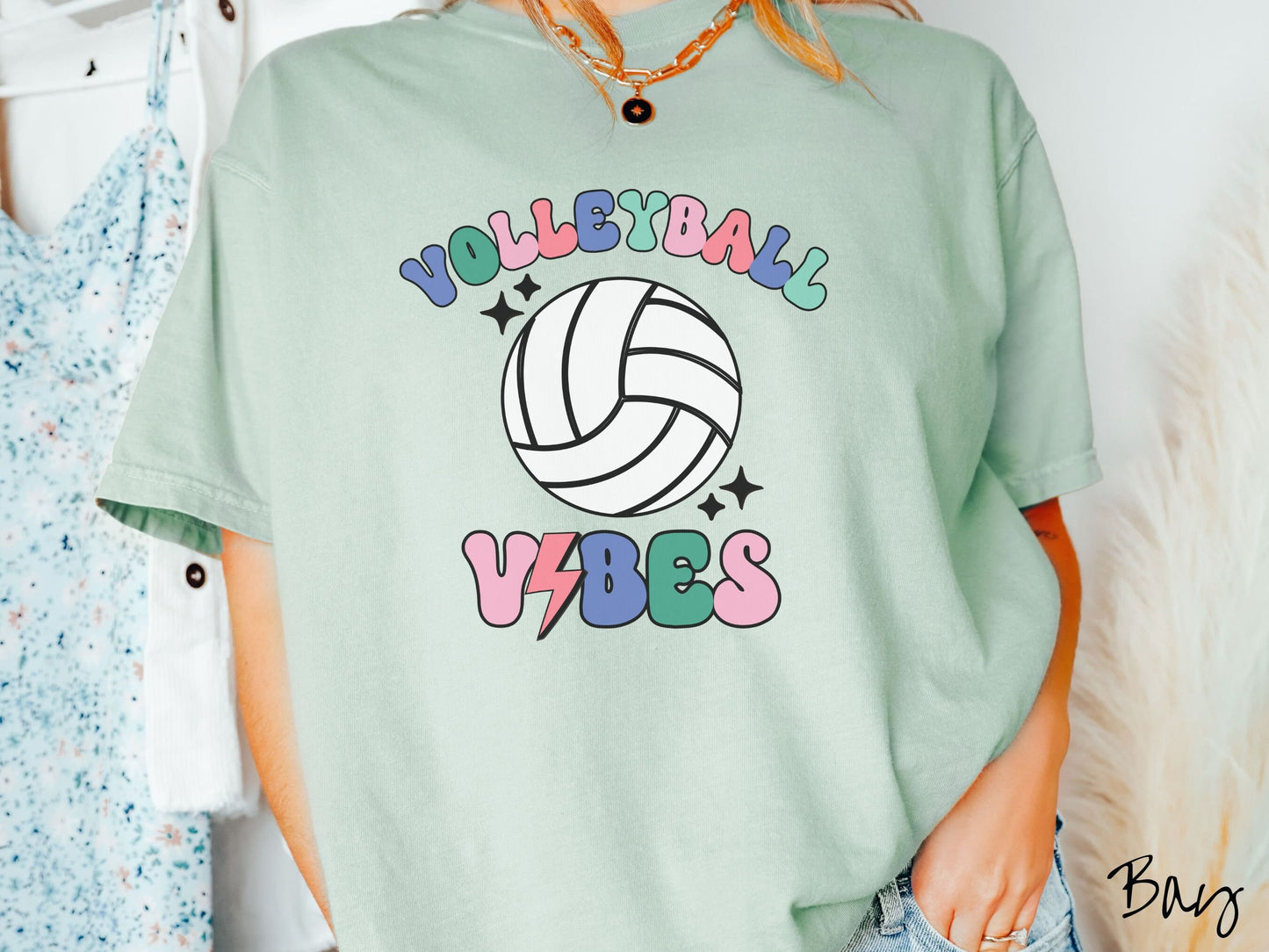 A woman wearing a cute, vintage bay colored Comfort Colors shirt with the word Volleyball across the front in colorful, uppercase fonts. Below that is a white volleyball and below that is the word vibes in colorful, uppercase fonts.