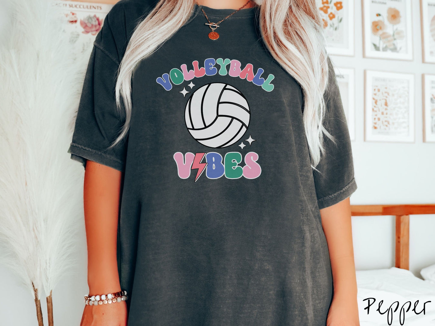 A woman wearing a cute, vintage pepper colored Comfort Colors shirt with the word Volleyball across the front in colorful, uppercase fonts. Below that is a white volleyball and below that is the word vibes in colorful, uppercase fonts.
