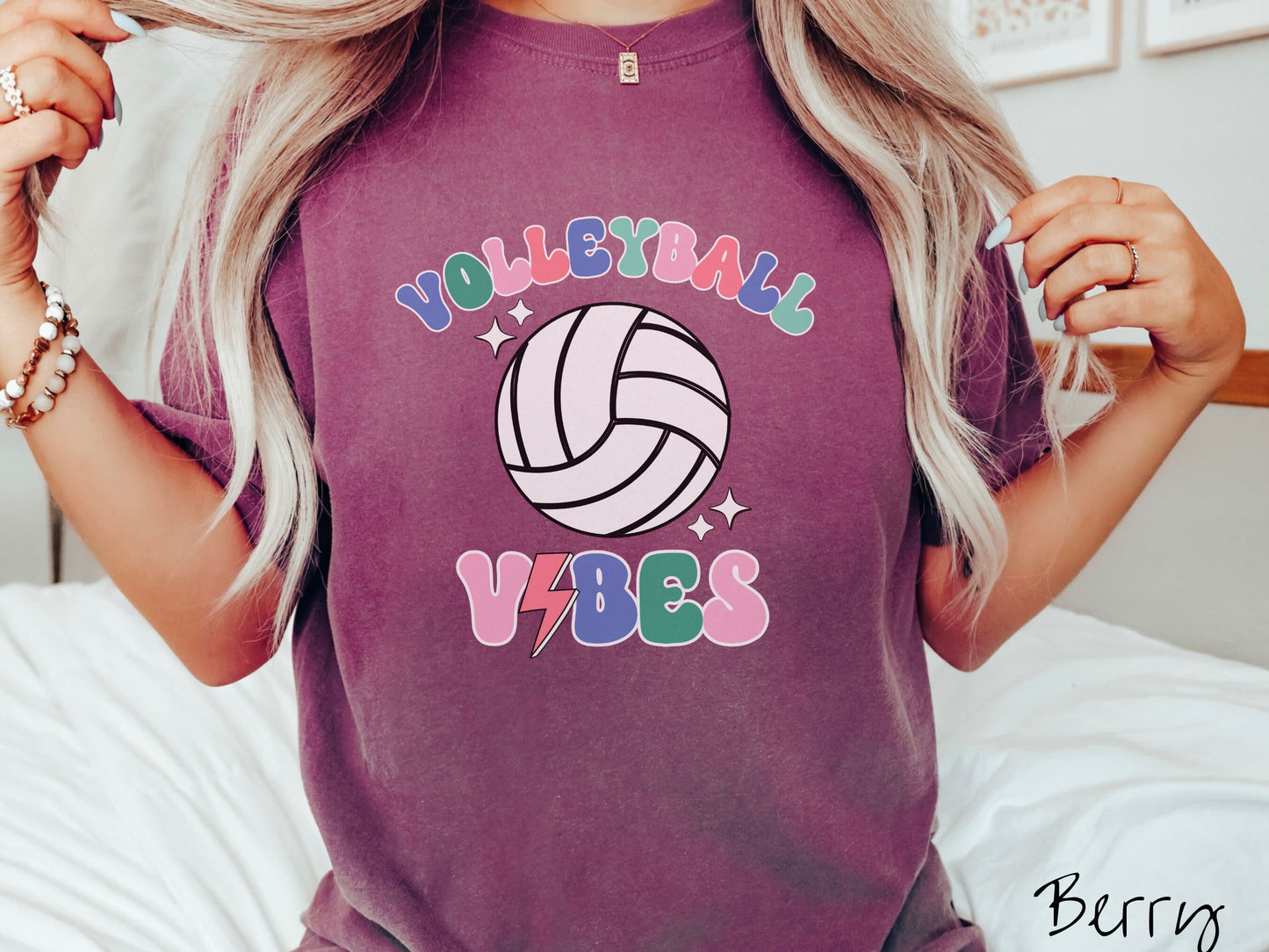 A woman wearing a cute, vintage berry colored Comfort Colors shirt with the word Volleyball across the front in colorful, uppercase fonts. Below that is a white volleyball and below that is the word vibes in colorful, uppercase fonts.