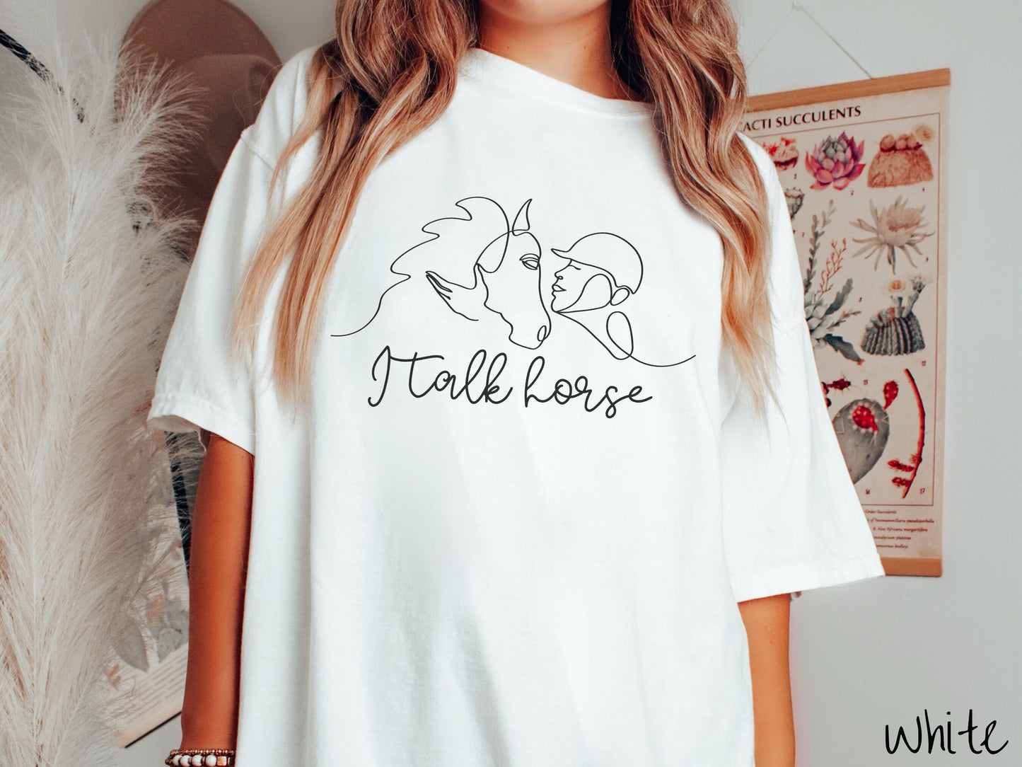 A woman wearing a cute, vintage white colored Comfort Colors shirt with a black silhouette outline of a horse jockey embracing his horse. Below that are the words I talk horse in black, cursive font.