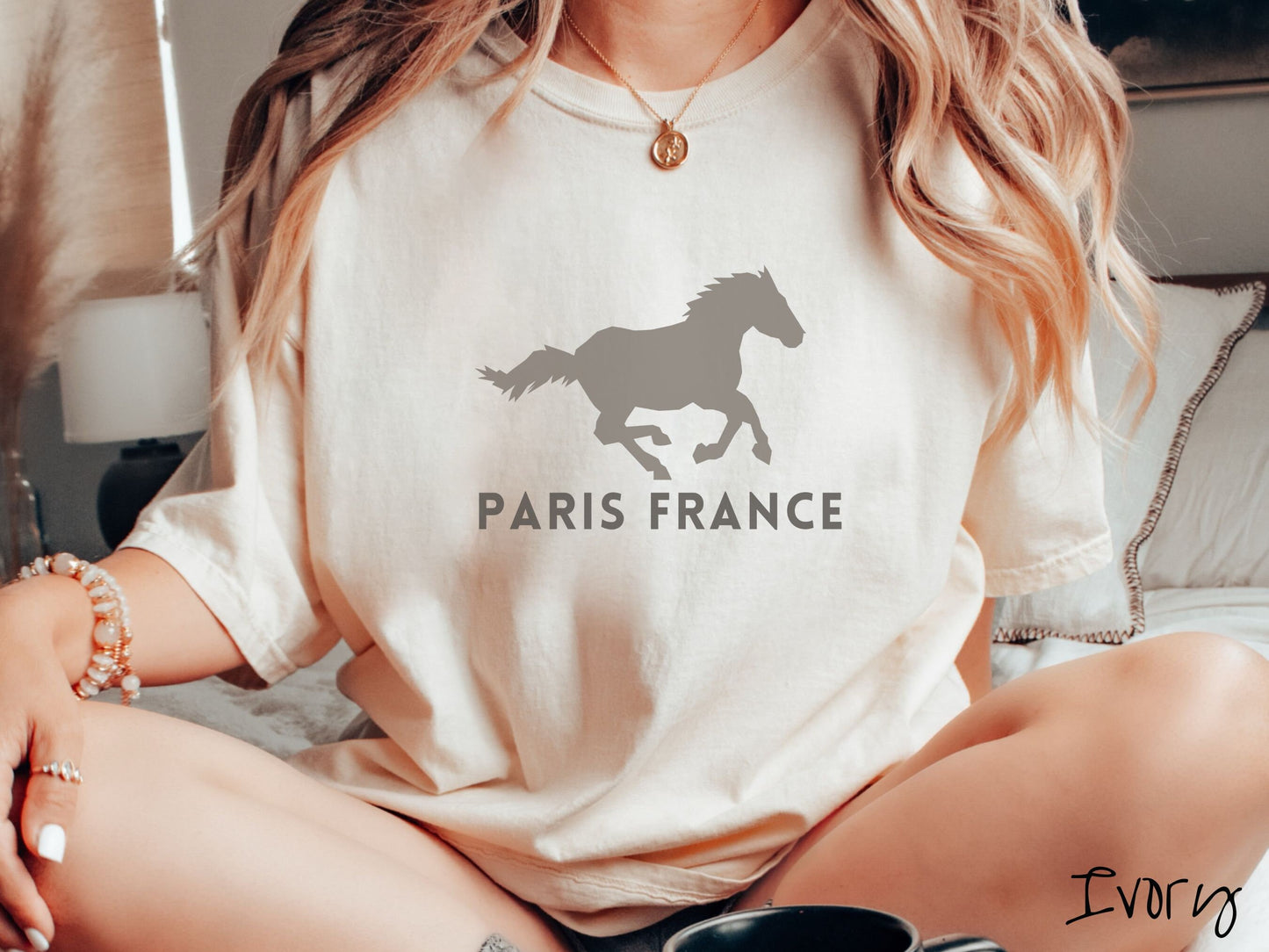 A woman wearing a cute, vintage ivory colored Comfort Colors shirt with a gray silhouette of a running horse across the font. Below it are the words Paris France in gray, uppercase font.