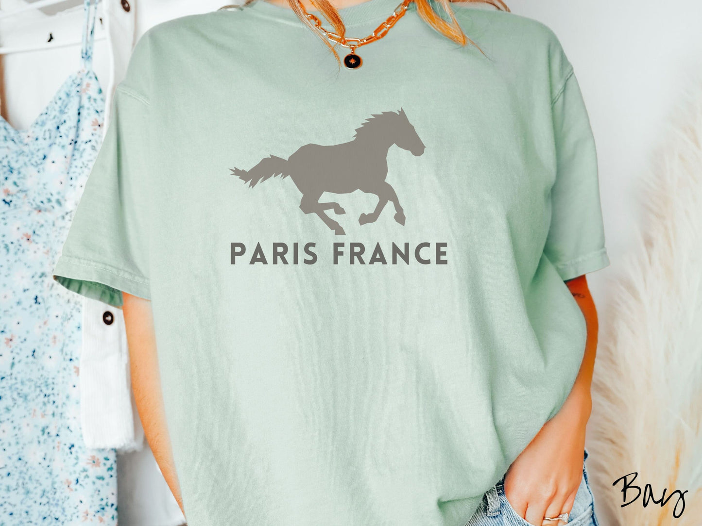 A woman wearing a cute, vintage bay colored Comfort Colors shirt with a gray silhouette of a running horse across the font. Below it are the words Paris France in gray, uppercase font.