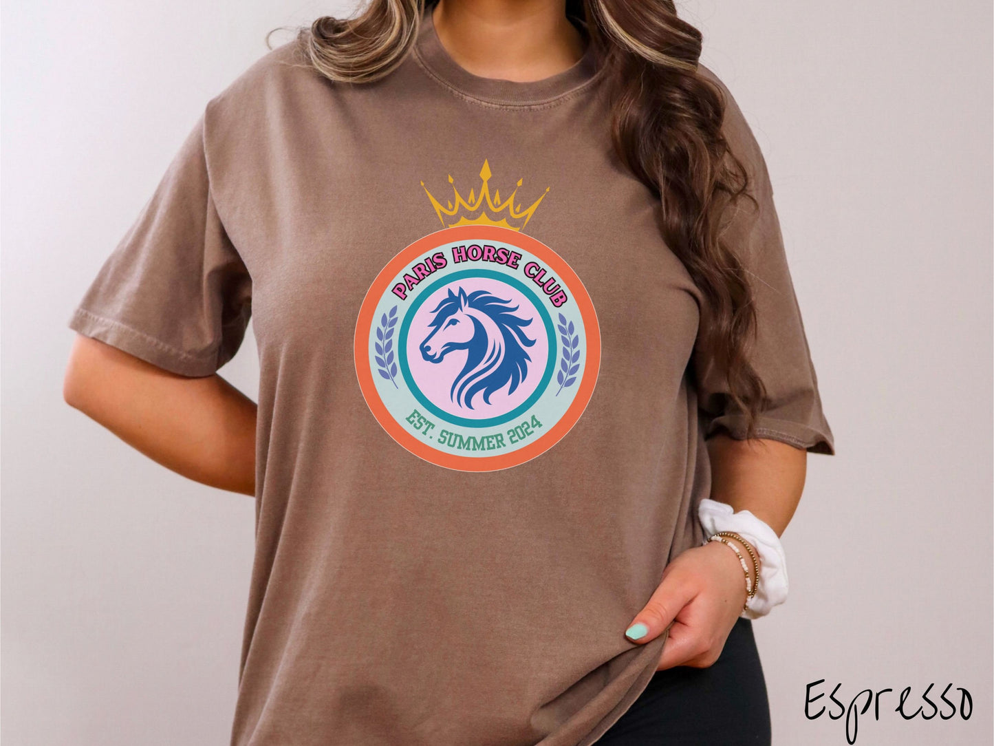 A woman wearing a cute, vintage espresso colored Comfort Colors shirt with a colorful coat of arms with a horse in the middle of two concentric circles, a crown on top. There is text along the top Paris Horse Club and below that Est. Summer 2024.