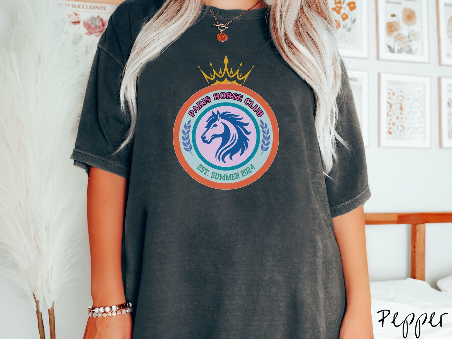 A woman wearing a cute, vintage pepper colored Comfort Colors shirt with a colorful coat of arms with a horse in the middle of two concentric circles, a crown on top. There is pink text along the top Paris Horse Club and below that Est. Summer 2024.