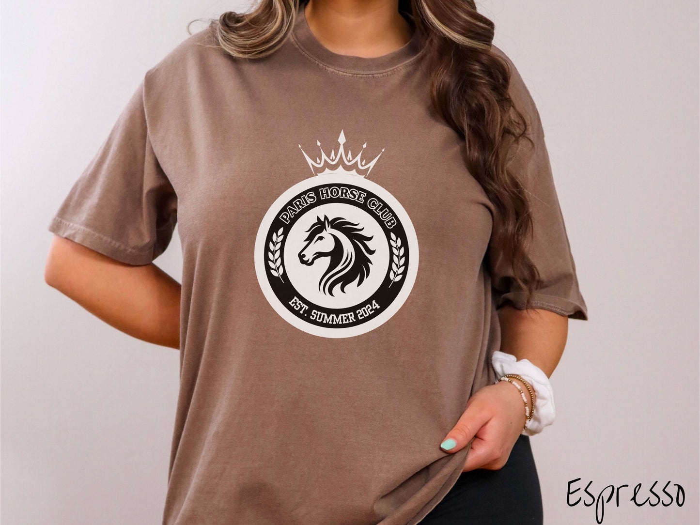 A woman wearing a cute, vintage espresso colored Comfort Colors shirt with a black and white coat of arms with a horse in the middle of two concentric circles, a crown on top. There is text along the top Paris Horse Club and below Est. Summer 2024.