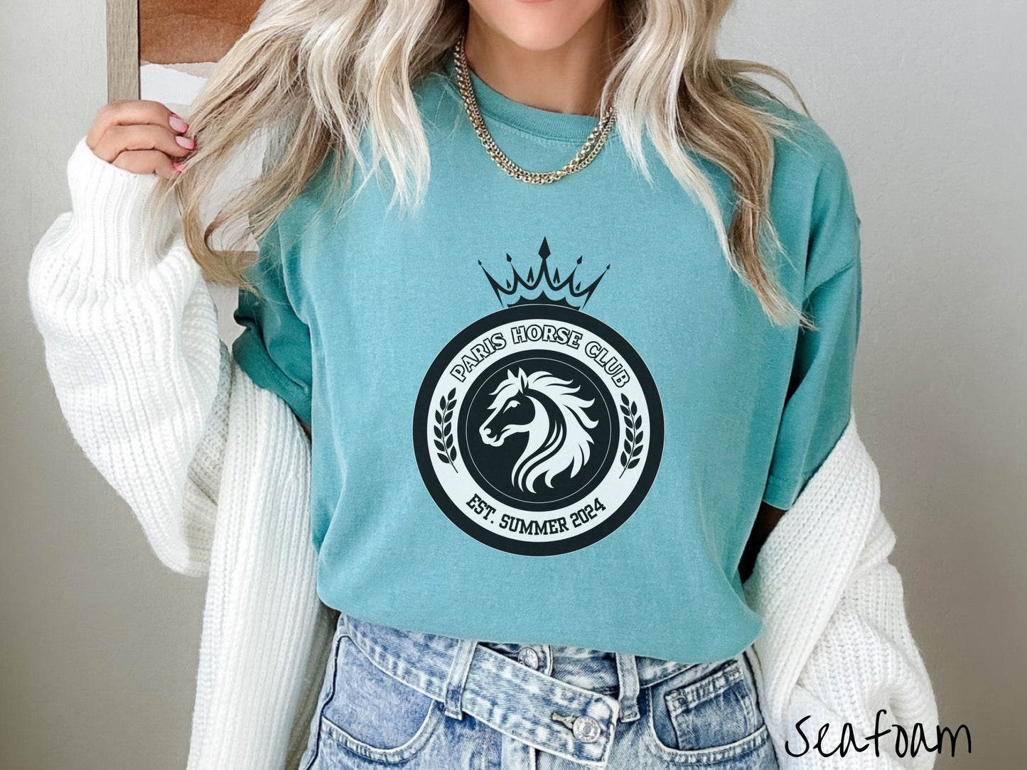 A woman wearing a cute, vintage seafoam colored Comfort Colors shirt with a black and white coat of arms with a horse in the middle of two concentric circles, a crown on top. There is text along the top Paris Horse Club and below Est. Summer 2024.
