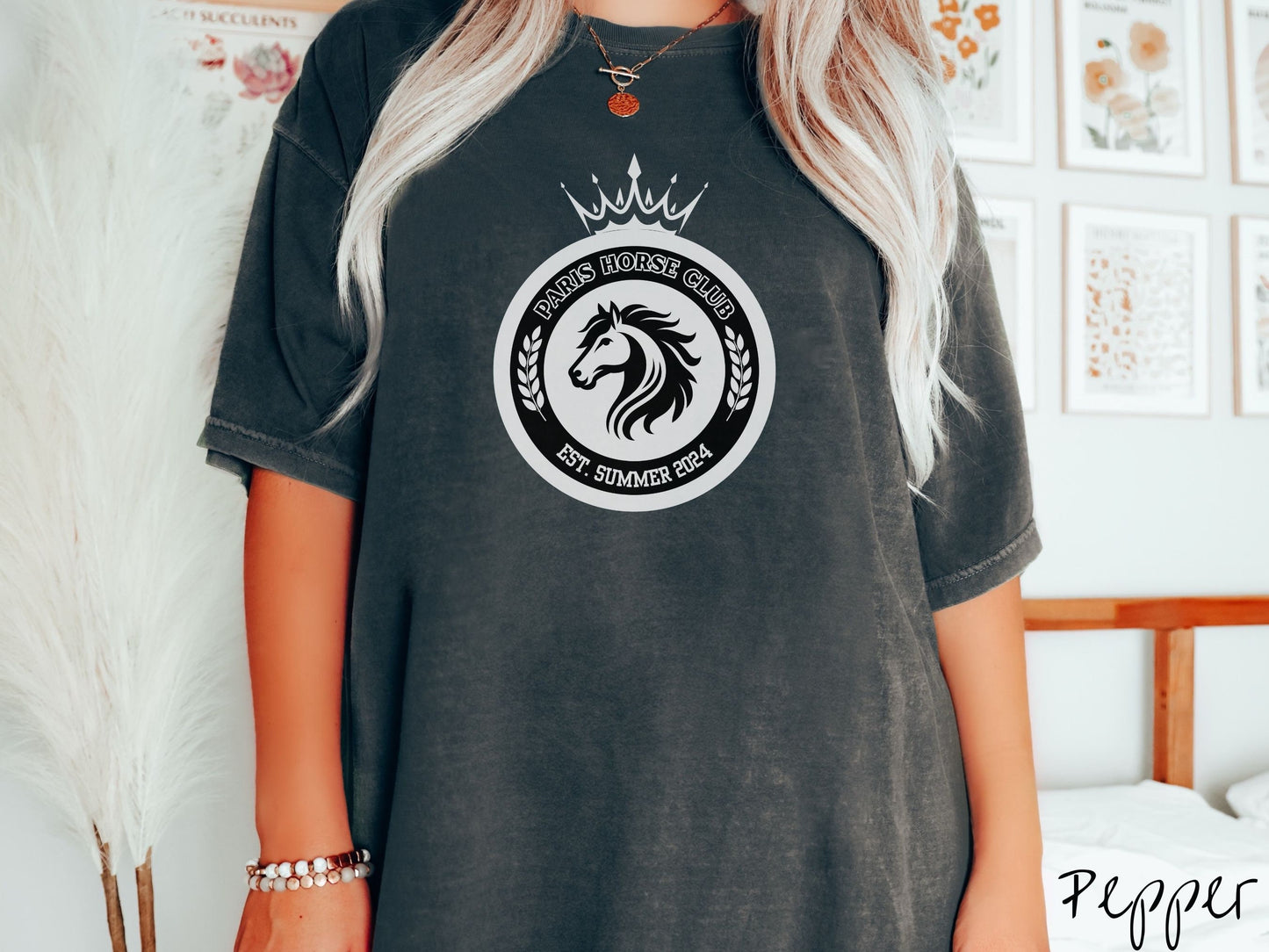 A woman wearing a cute, vintage pepper colored Comfort Colors shirt with a black and white coat of arms with a horse in the middle of two concentric circles, a crown on top. There is text along the top Paris Horse Club and below Est. Summer 2024.
