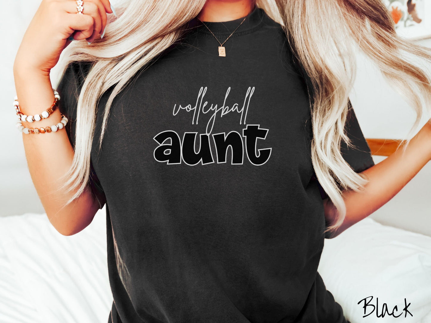 A woman wearing a cute, vintage black colored Comfort Colors shirt with the word volleyball across the front in white, undercase cursive writing. Below that is the word aunt in large, black font.