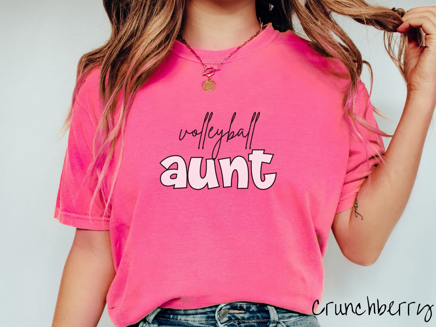 A woman wearing a cute, vintage crunchberry colored Comfort Colors shirt with the word volleyball across the front in black, undercase cursive writing. Below that is the word aunt in large, light pink font.