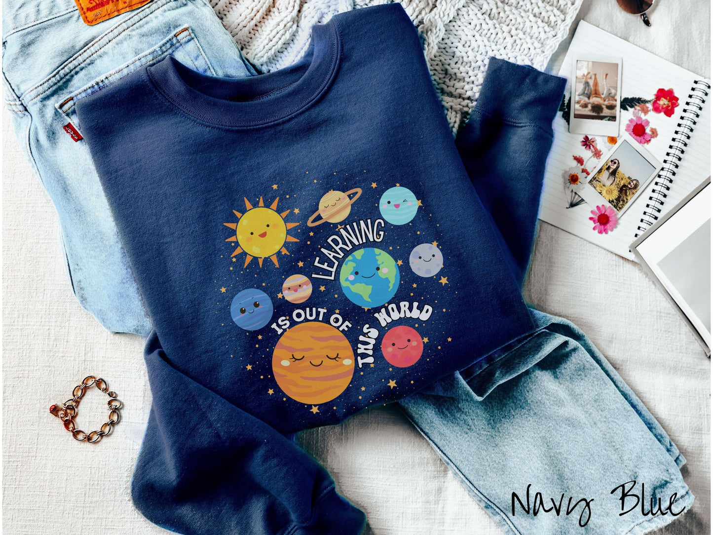 A cute, vintage navy blue colored sweatshirt showing the solar system and the planets have smiling, happy faces. There is text saying Learning Is Out of This World among the planets, and there are many stars twinkling in the background.