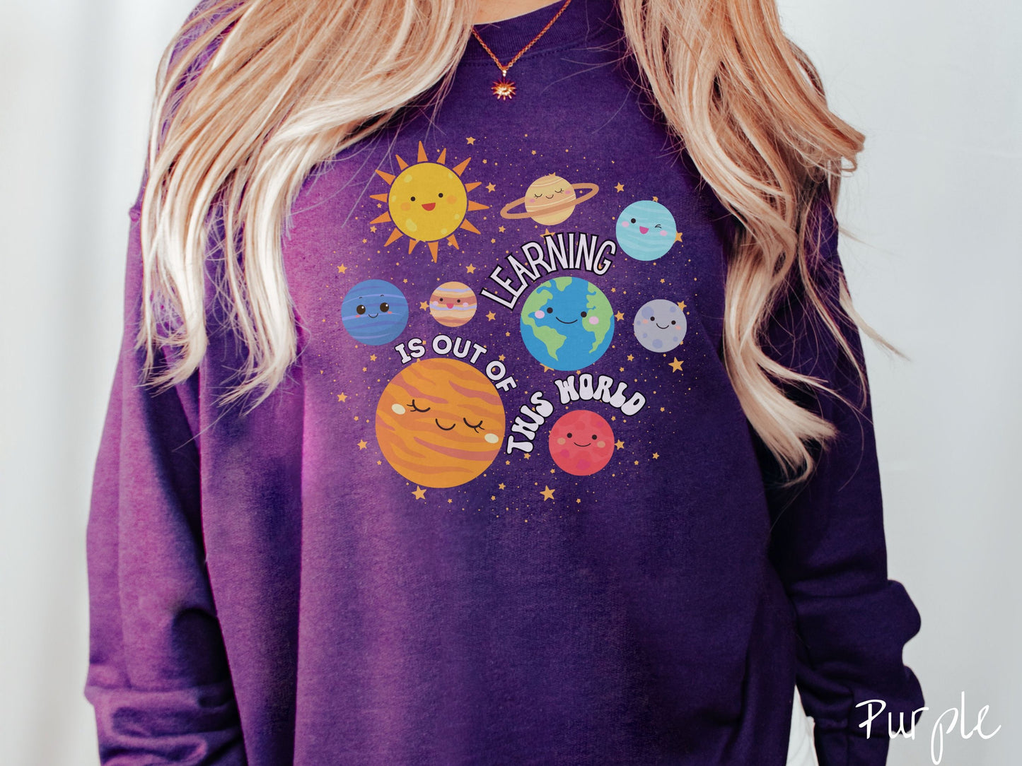 A woman wearing a cute, vintage purple colored sweatshirt showing the solar system and the planets have smiling, happy faces. There is text saying Learning Is Out of This World among the planets, and there are many stars twinkling in the background.