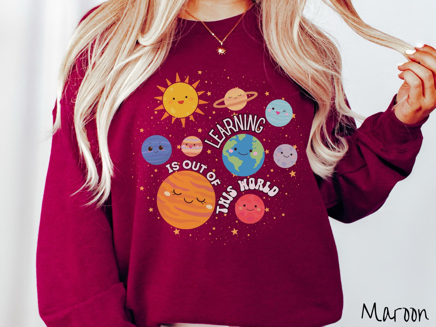 A woman wearing a cute, vintage red colored sweatshirt showing the solar system and the planets have smiling, happy faces. There is text saying Learning Is Out of This World among the planets, and there are many stars twinkling in the background.