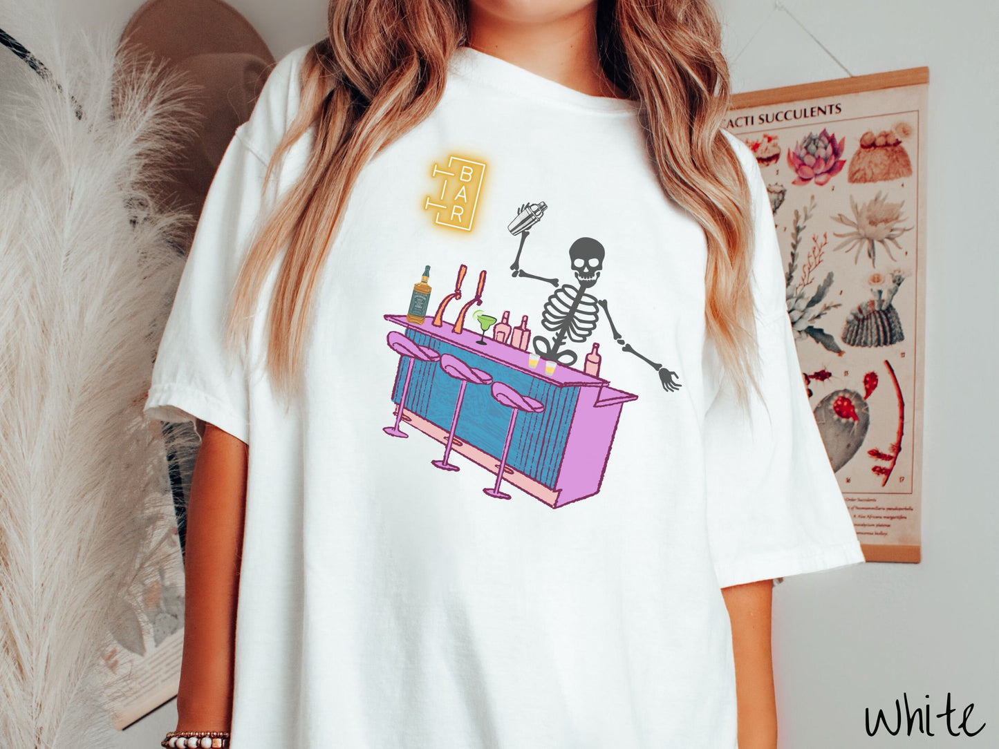 A woman wearing a cute, vintage white colored Comfort Colors t-shirt showing a skeleton bartender behind a purple and blue bar mixing drinks. There is a yellow bar sign behind the bar and three purple chairs in front. There are liquor bottles on top.
