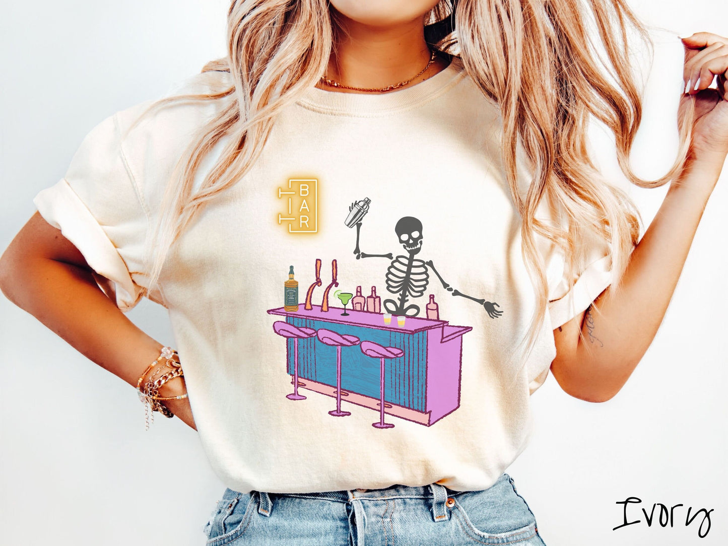 A woman wearing a cute, vintage ivory colored Comfort Colors t-shirt showing a skeleton bartender behind a purple bar mixing drinks. There is a yellow bar sign behind the bar and three purple chairs in front. There are liquor bottles on top.